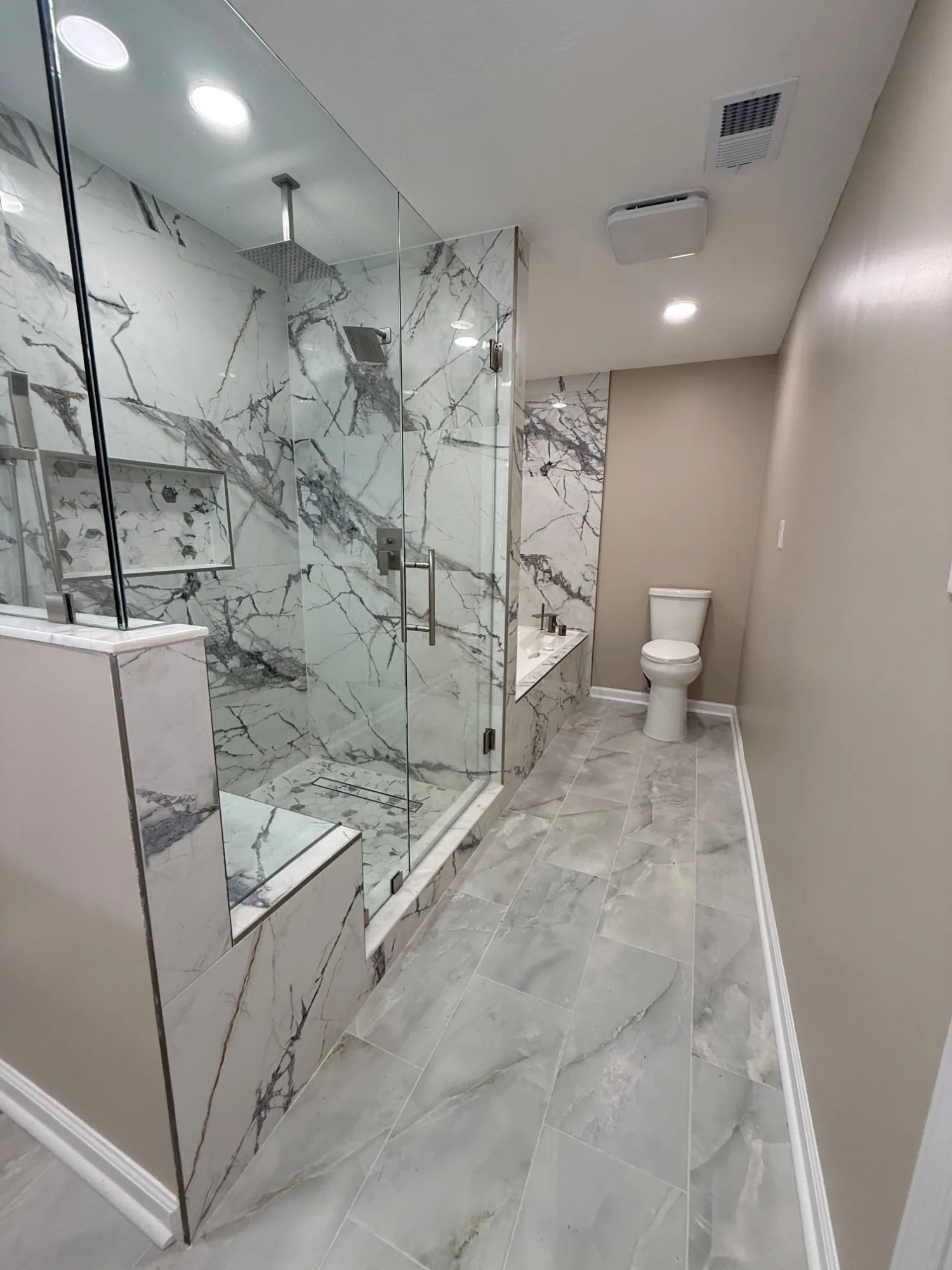 The Richmond Hill primary bathroom: a walk-in shower behind frameless glass on a stone-capped half wall, marble-look tile floor to ceiling, and a soaking tub set into a tiled deck beyond it.