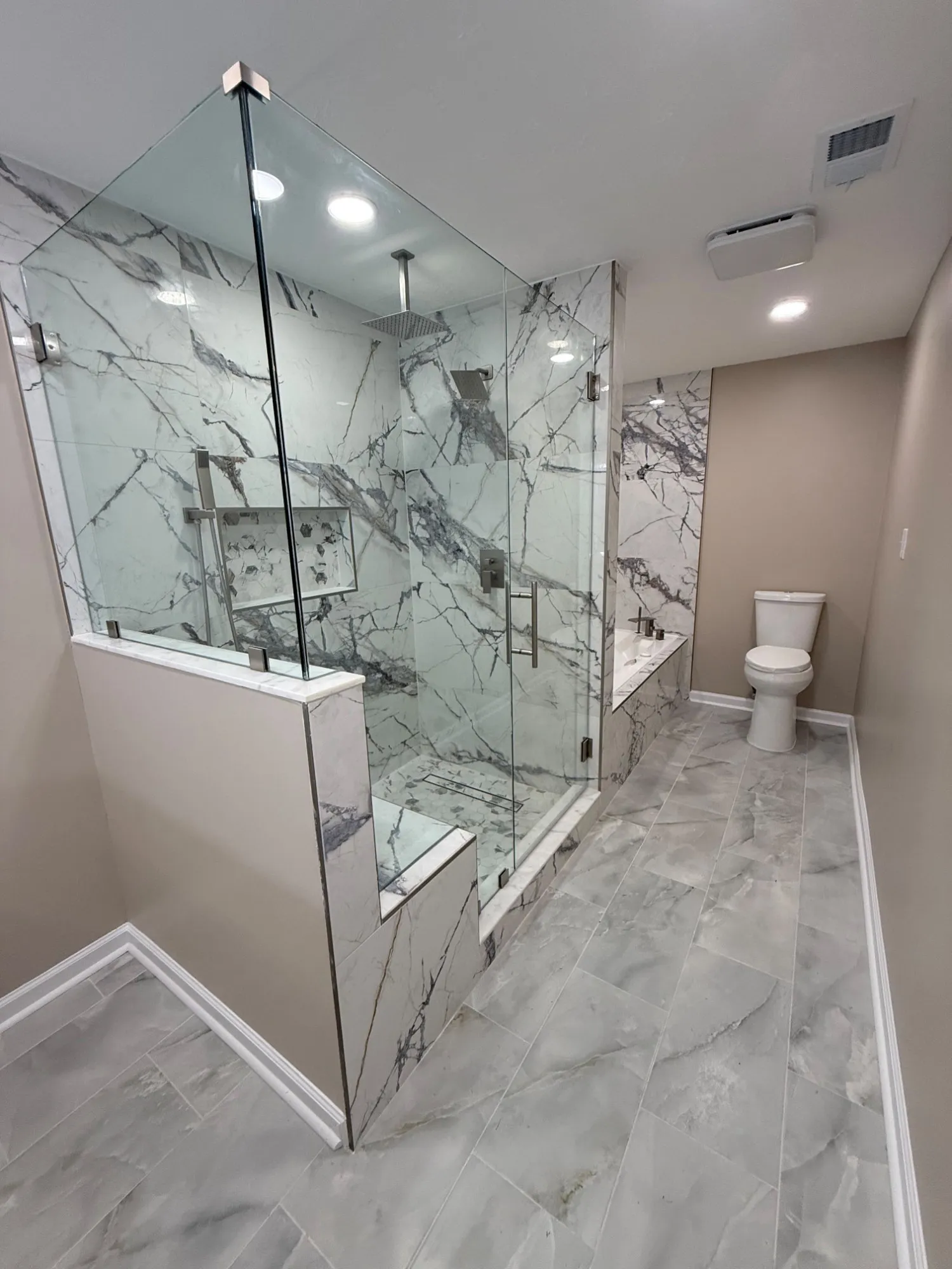 The primary bathroom from the doorway: the glass shower enclosure, the tub deck and the large-format tile floor under recessed lighting.