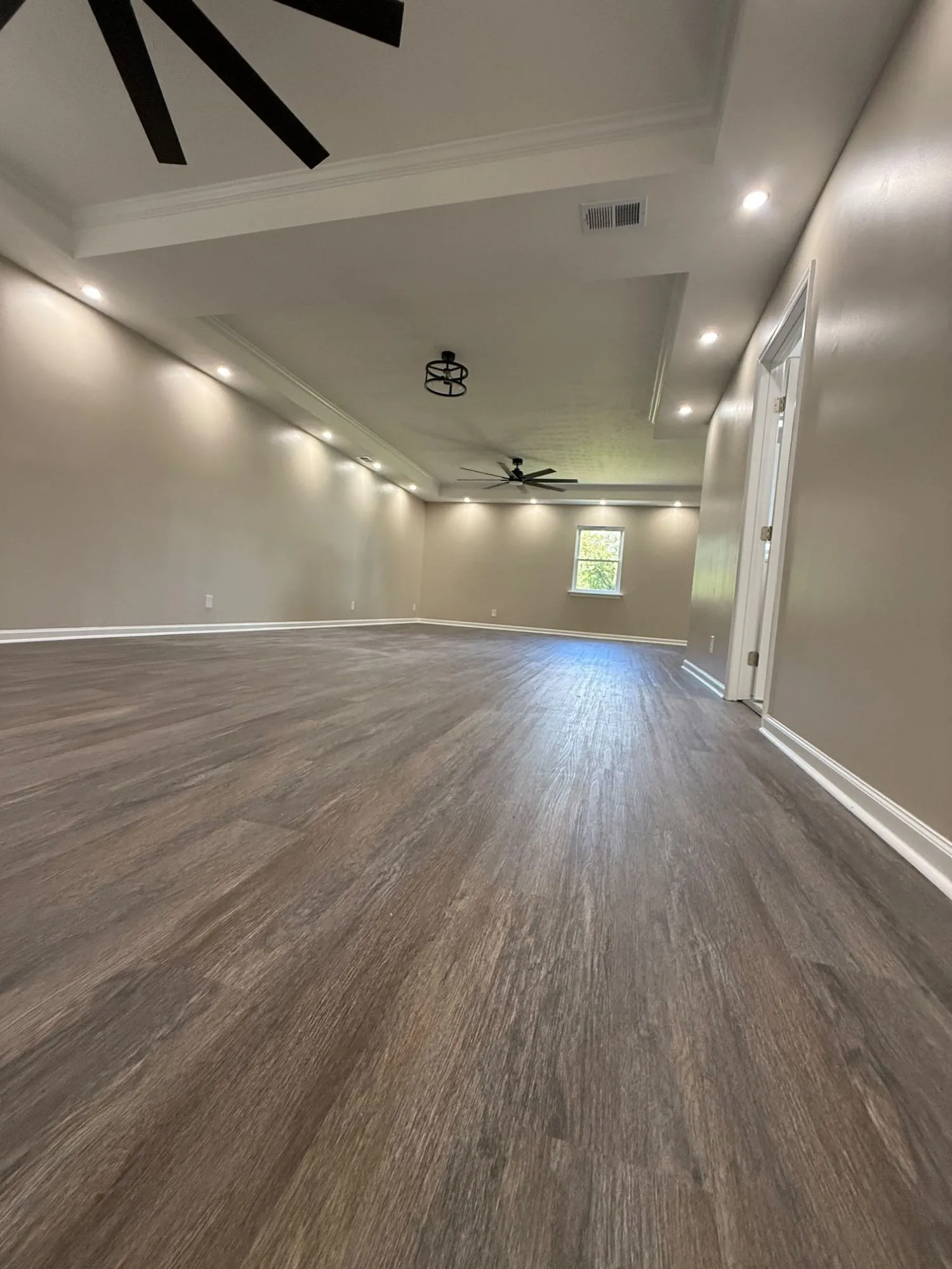 The primary bedroom after the renovation: wide-plank flooring wall to wall, a tray ceiling with crown and recessed lighting, and a new ceiling fan.