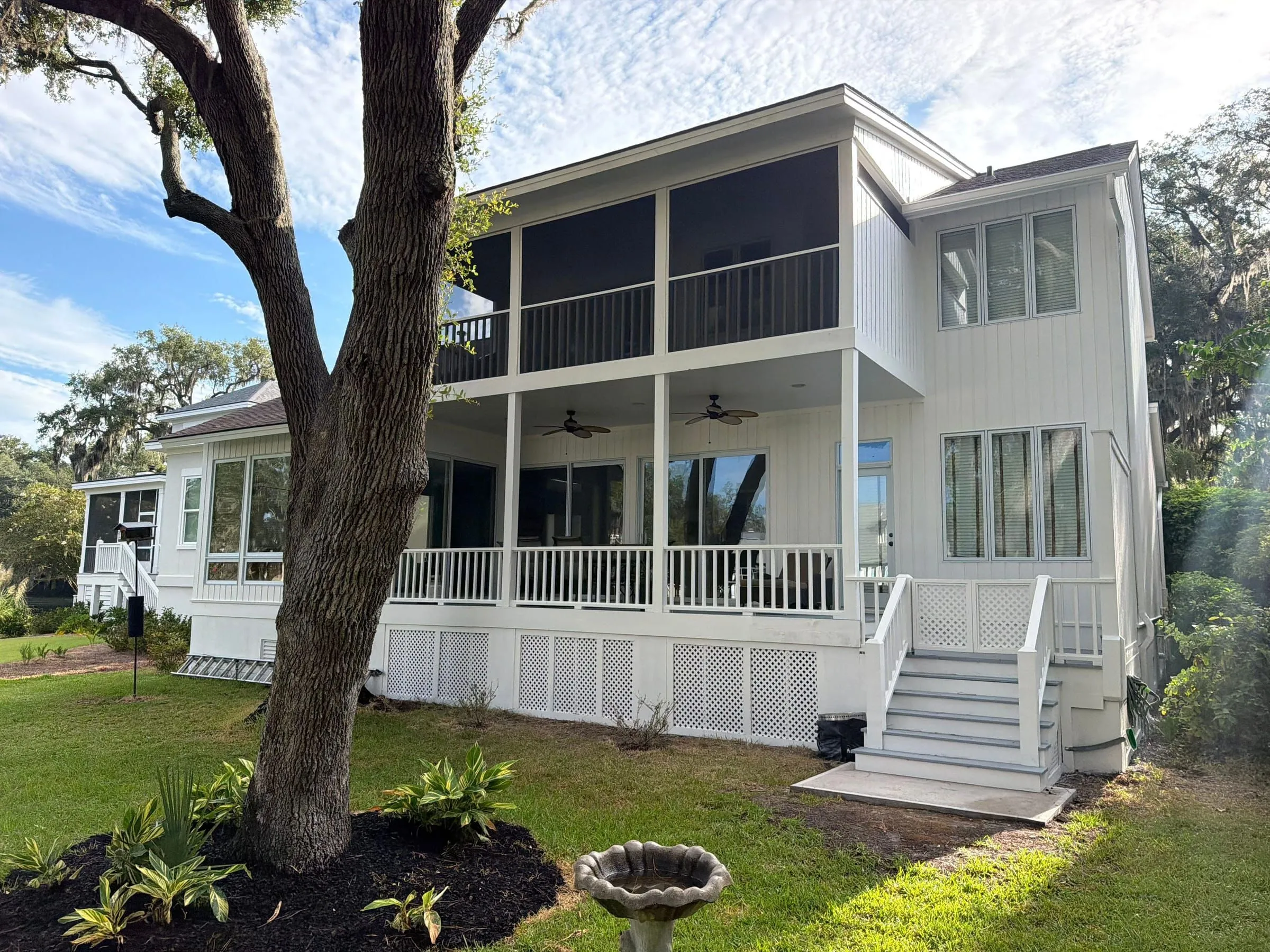 A two-story white board-and-batten house under live oaks, screened porch above and a railed porch below, with new steps down to the lawn.