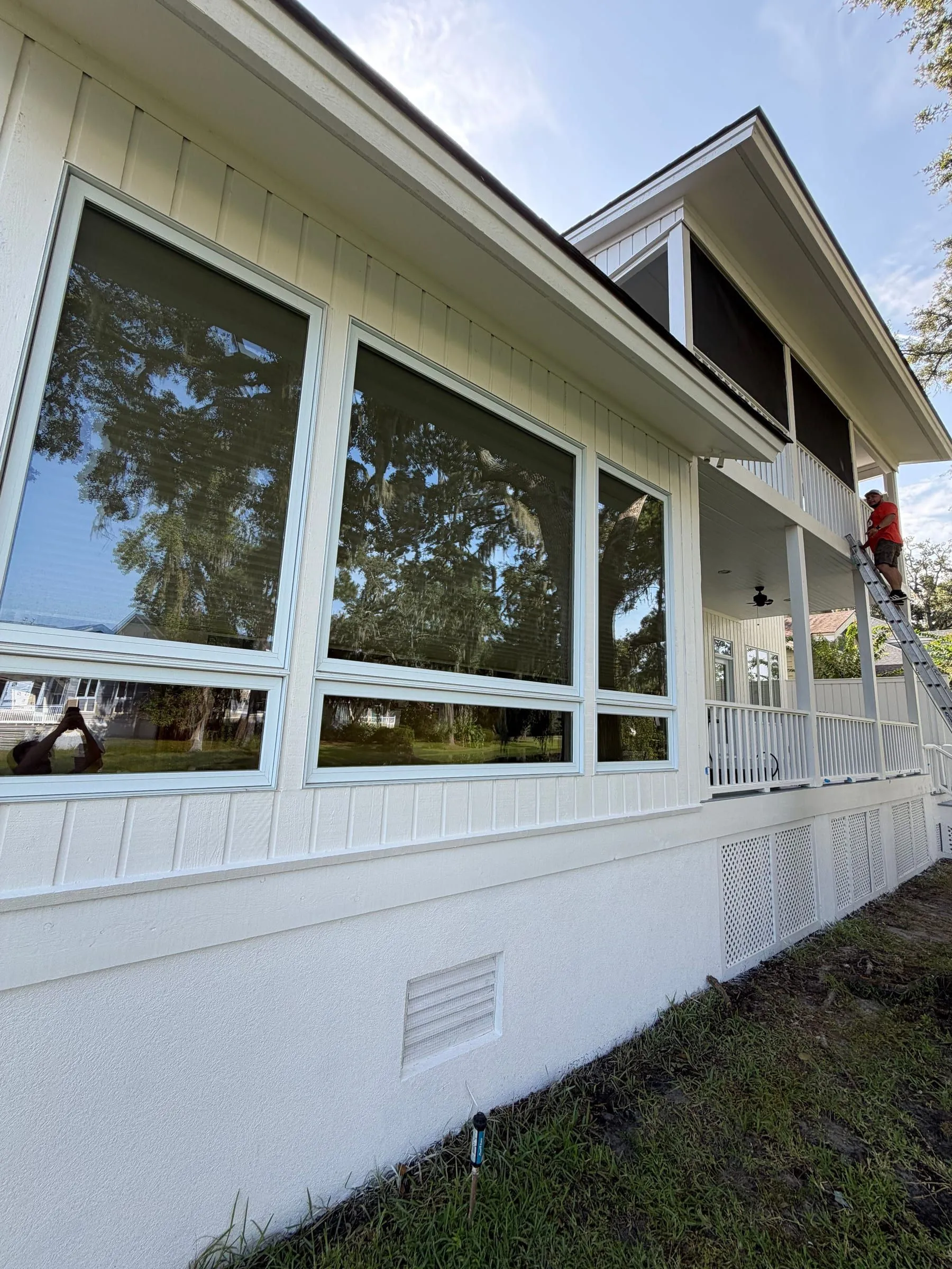 A white weatherboard elevation seen from below: eave and gutter line, board-and-batten siding, a wall of windows reflecting live oaks, and a screened porch beyond.
