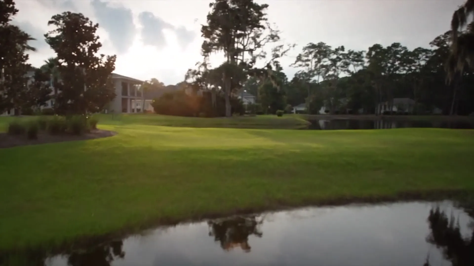 Late afternoon light across a lawn and lagoon, live oaks and houses beyond. A coastal Georgia house at sunset, its windows lit above the water and marsh.
