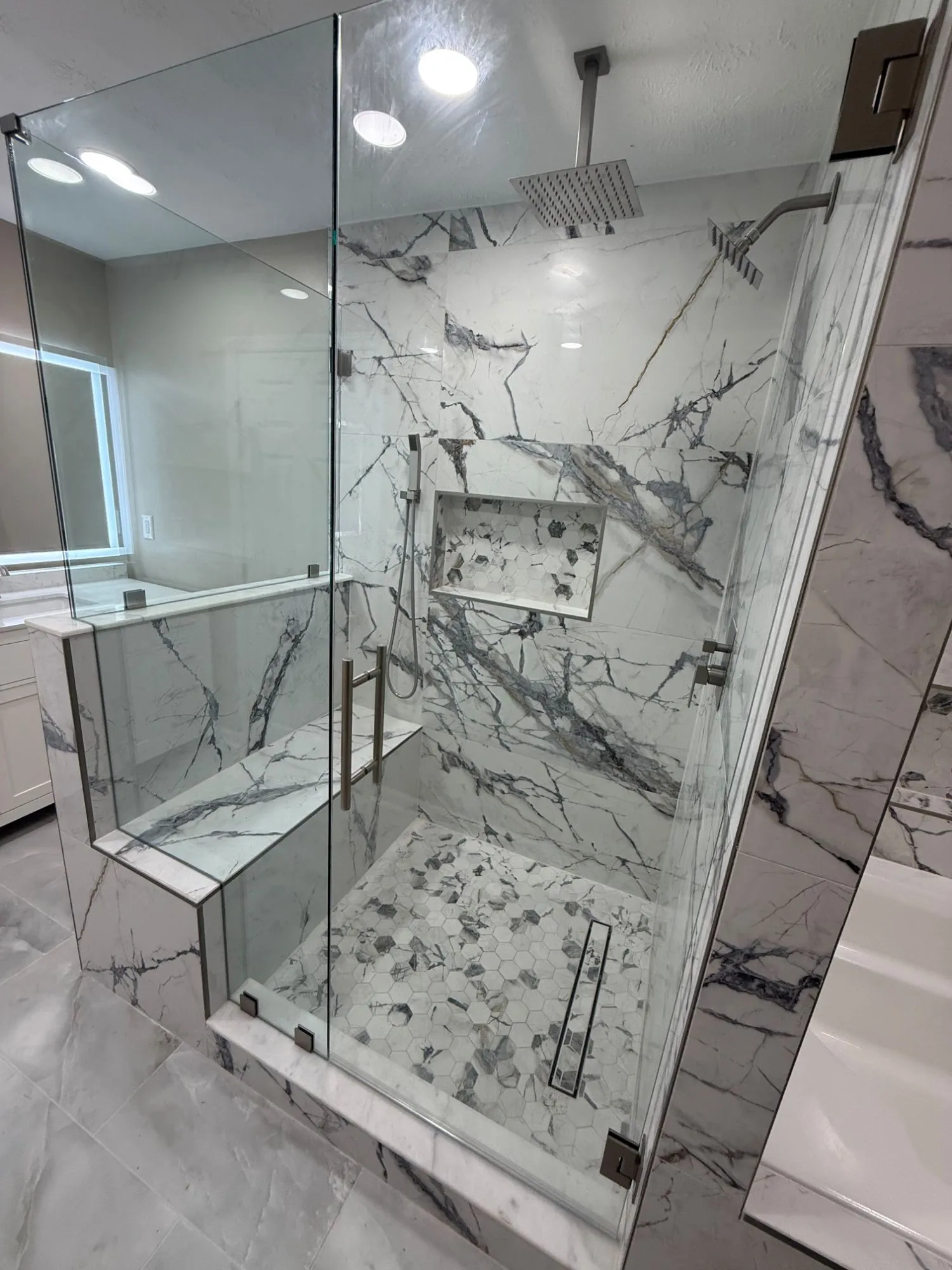 Inside the walk-in shower: a rain head from the ceiling, a second head on the wall, a hand shower on a slide bar, a bench, a niche lined in hexagon mosaic and the same mosaic underfoot running to a linear drain.